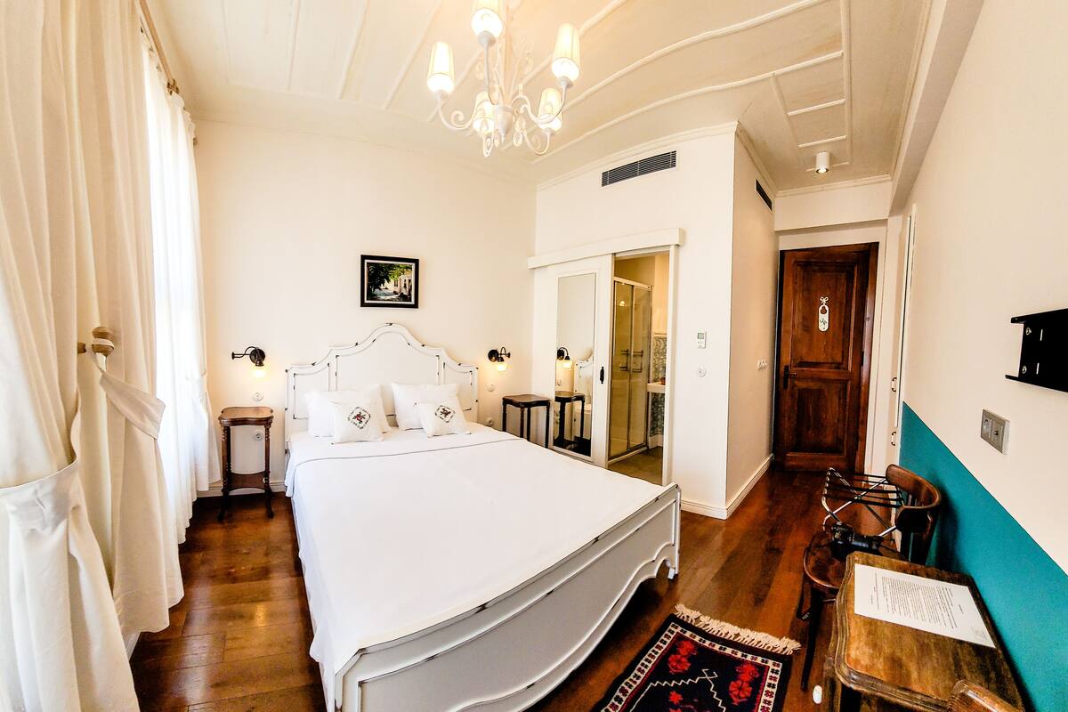 A bedroom with a large bed, a chandelier, a bedside table, a nightstand, a wooden floor, and a door leading to a bathroom.