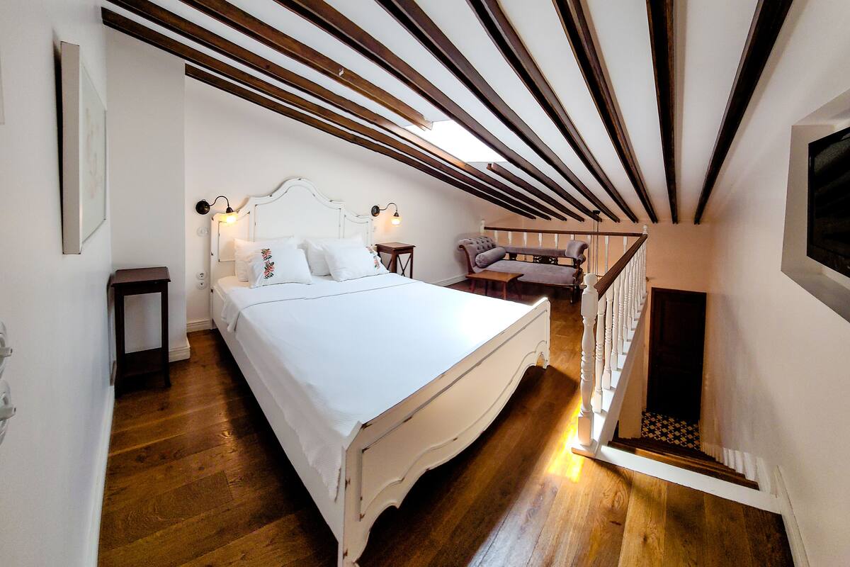 A bedroom with a large bed, wooden beams on the ceiling, a small table, and a staircase leading to another room.