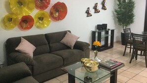 Un saló amb un sofà, una taula de centre de vidre, plats decoratius amb flors, una petita planta i una zona de menjador amb cadires.