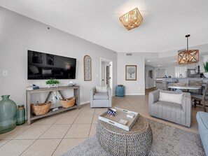 A modern living room with a flat-screen TV, grey sofas, a coffee table, and a dining area with a kitchen in the background.
