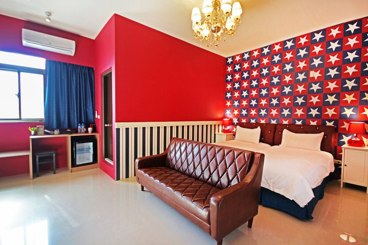 A hotel room with a red and white star-patterned wall, a brown leather sofa, a bed with white bedding, a chandelier, and a small desk with a refrigerator.