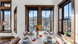 A dining room with a wooden table set for four, large windows with mountain views, and a vase of flowers on the table. - Aurum Condo -1 BR w/ Mtn Views, Steps from Main St (Breckenridge)