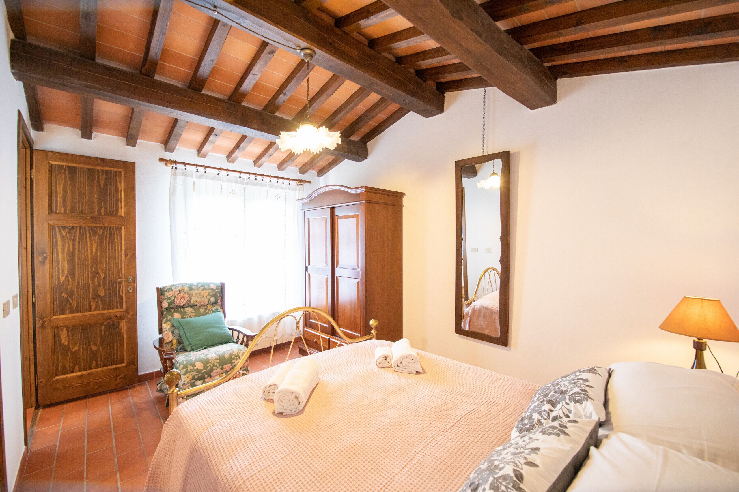 Quarto com teto de madeira, cama com toalhas dobradas, cadeira com estofamento floral e um espelho na parede.