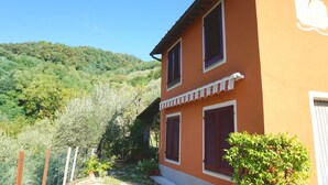 A two-story building with a sloped roof, a balcony with a striped awning, and a view of a lush green hillside.