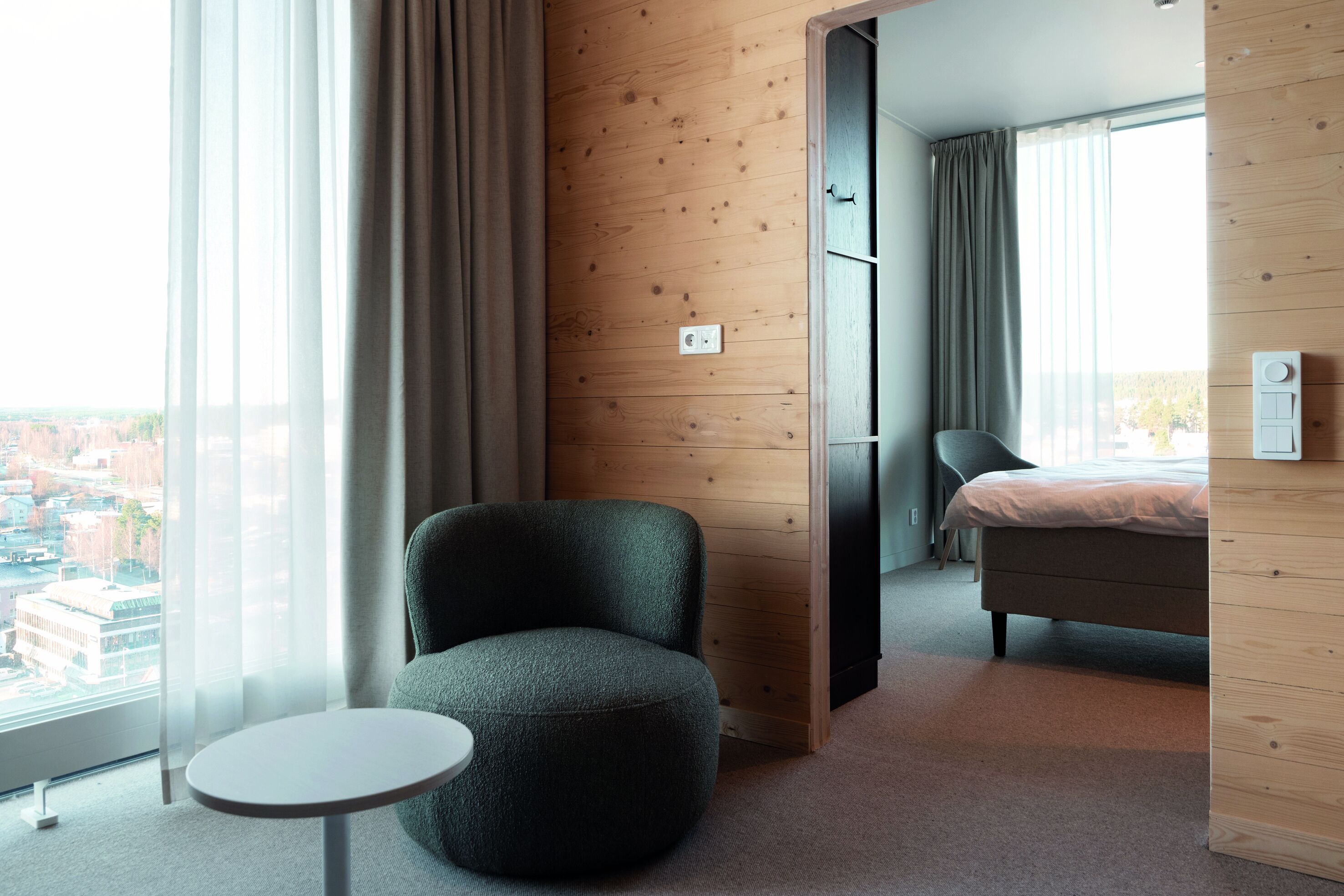 Une chambre d’hôtel moderne avec un pouf rond, un lit et une vue sur la ville grâce à de grandes fenêtres.