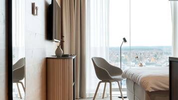 A modern hotel room with a large window, a bed, a chair, a TV, and a bedside table.