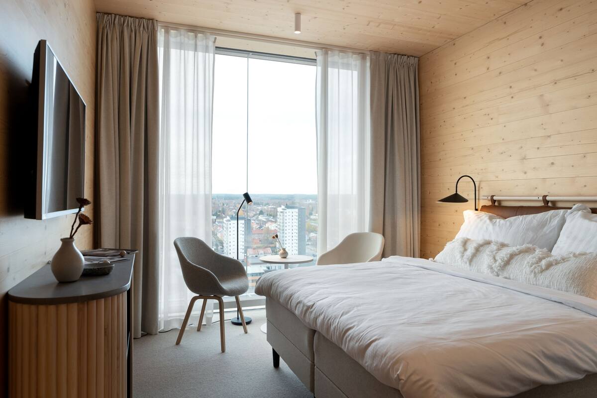 Une chambre d’hôtel avec un grand lit, un bureau agrémenté d’un vase, une chaise et une vue sur la ville grâce à de grandes fenêtres.