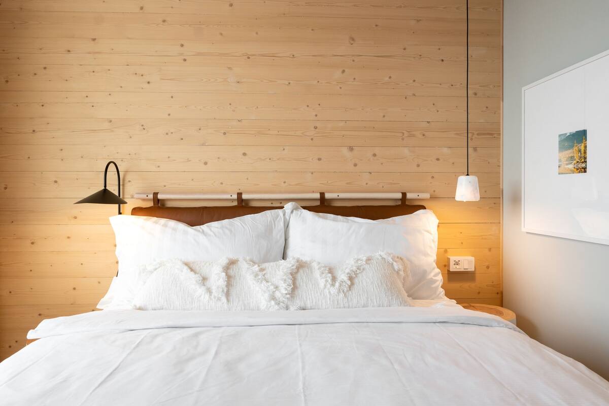 Un lit bien fait, avec du linge de lit blanc et des oreillers moelleux, une tête de lit en bois, une lampe de chevet noire et un luminaire suspendu.
