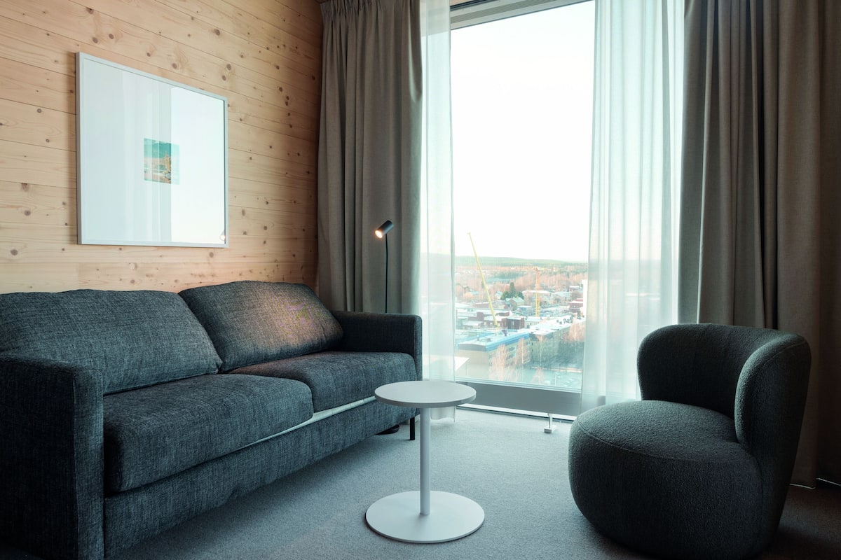 Un salon moderne avec un canapé gris, une table basse ronde blanche et un fauteuil foncé. Une grande fenêtre offre une vue sur le paysage urbain.