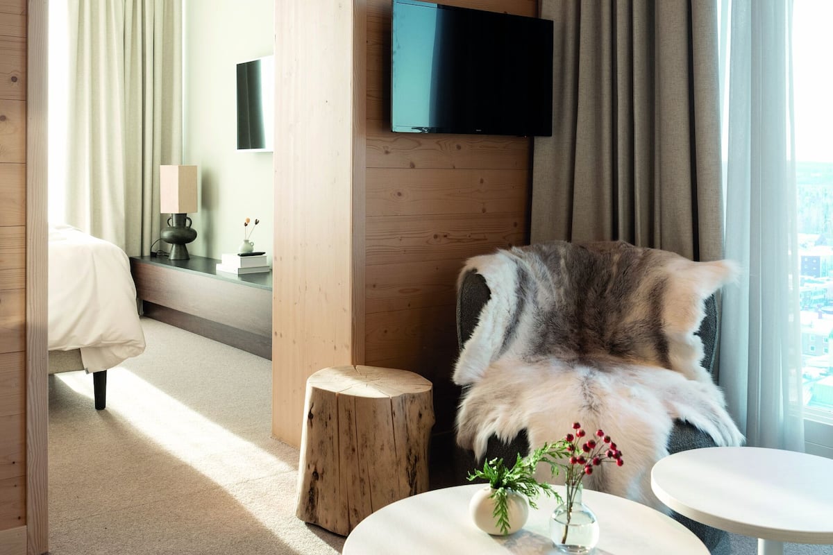 Une chambre d’hôtel moderne avec des murs en bois, un téléviseur à écran plat fixé au-dessus d’une fenêtre, un fauteuil moelleux et deux petites tables ornées d’objets décoratifs.
