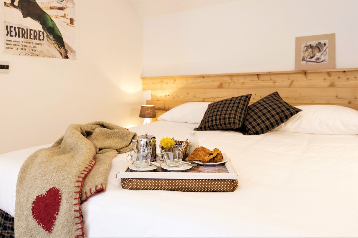 Une chambre d’hôtel avec une tête de lit en bois, un lit avec des oreillers blancs et à carreaux, un plateau avec un croissant et une théière, et une couverture ornée d’un motif en forme de cœur.