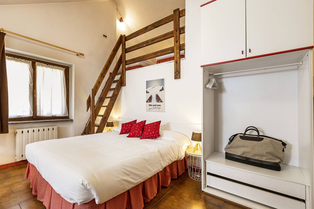 Un dormitorio con cama, escalera de madera, radiador, ventana con cortinas, un cuadro colgado en la pared y una estantería con un bolso.