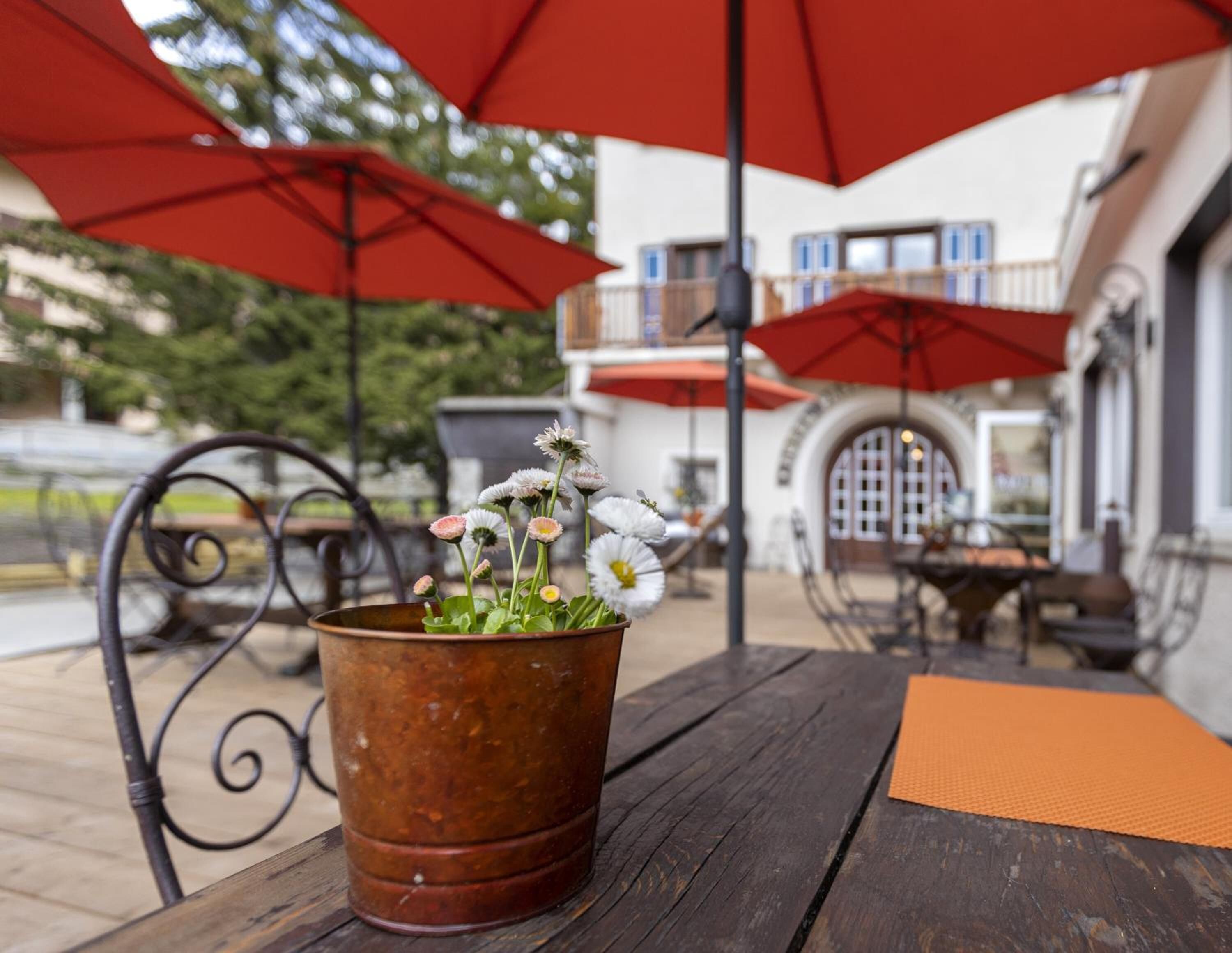 Ein rustikaler Tisch im Freien mit einem Metalltopf voller Blumen, roten Sonnenschirmen und einem Gebäude mit Rundbogenfenstern im Hintergrund.