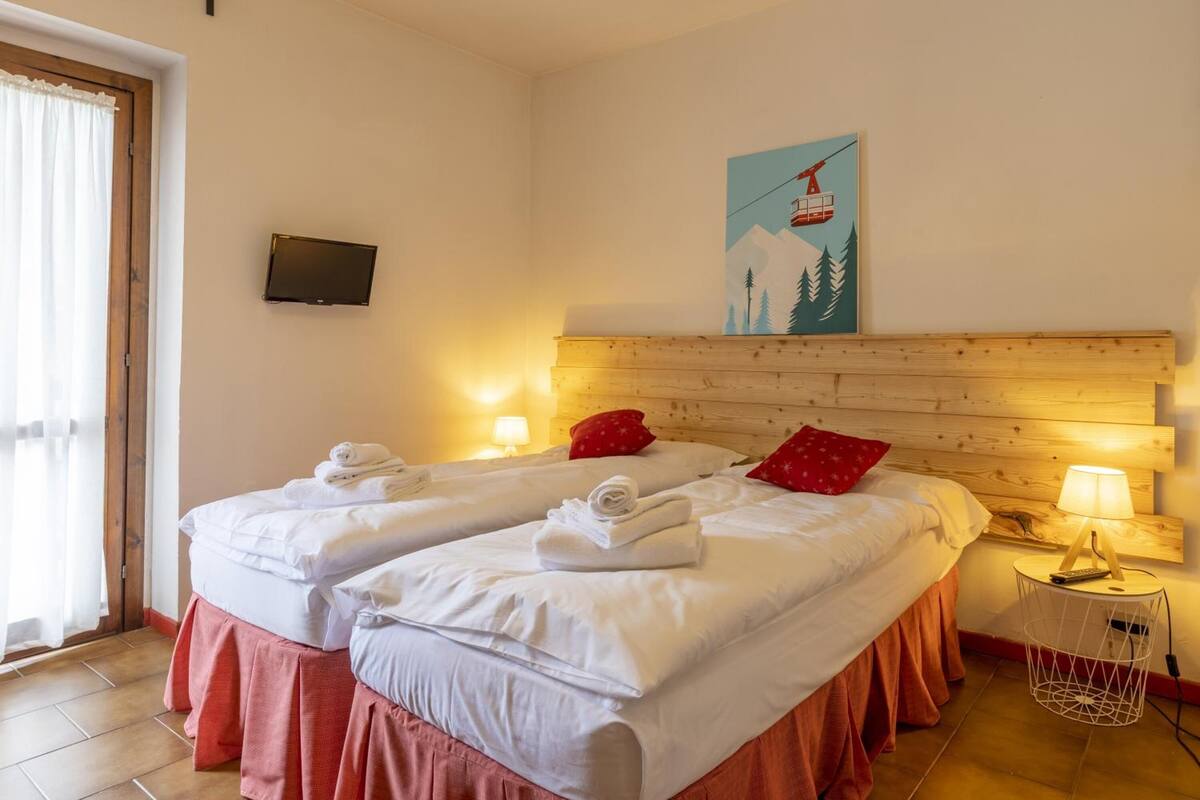 Habitación de hotel con dos camas, cabecera de madera, un cuadro con paisaje de montaña, televisor montado en la pared y una mesita de noche con lámpara y estación de carga.