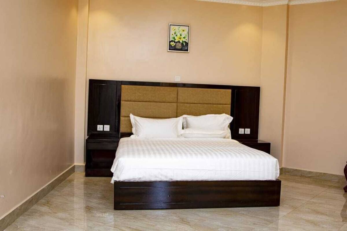 Quarto de hotel com uma cama grande, uma mesinha de cabeceira, um pequeno banco de madeira e um quadro na parede.