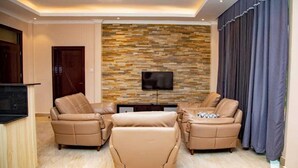 Sala de estar con dos sofás beige, una mesa de centro, un televisor de pantalla plana montado en una pared con diseño de piedra y una cortina oscura a la derecha.