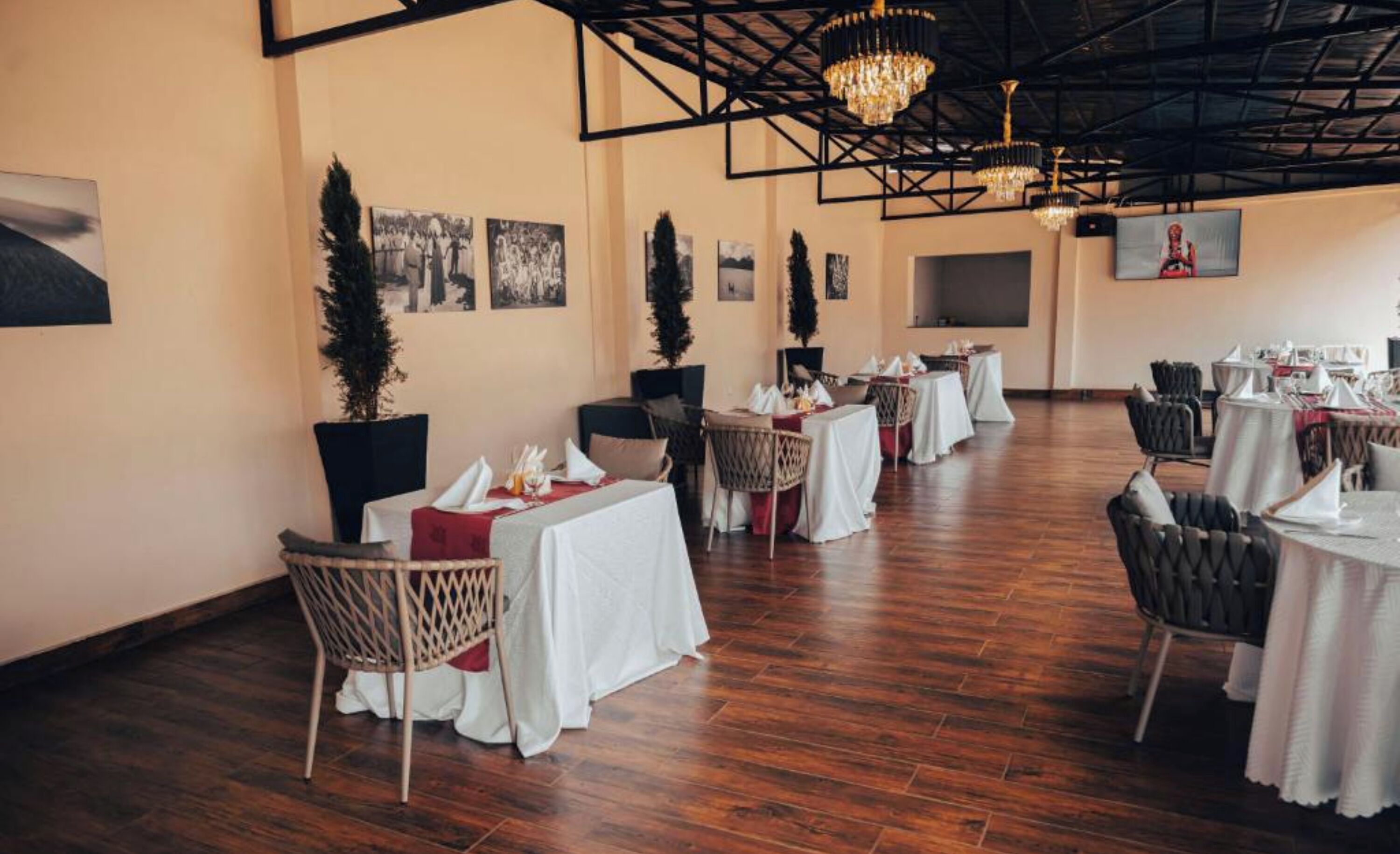 A spacious dining area with high ceilings, chandeliers, and a wooden floor. Tables are set with white tablecloths and chairs.