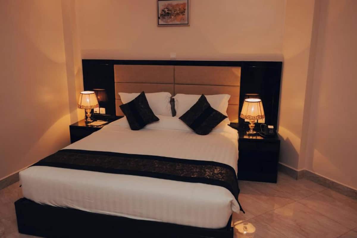Cama bem arrumada com cabeceira preta, duas mesas de cabeceira com luminárias e um quadro na parede.
