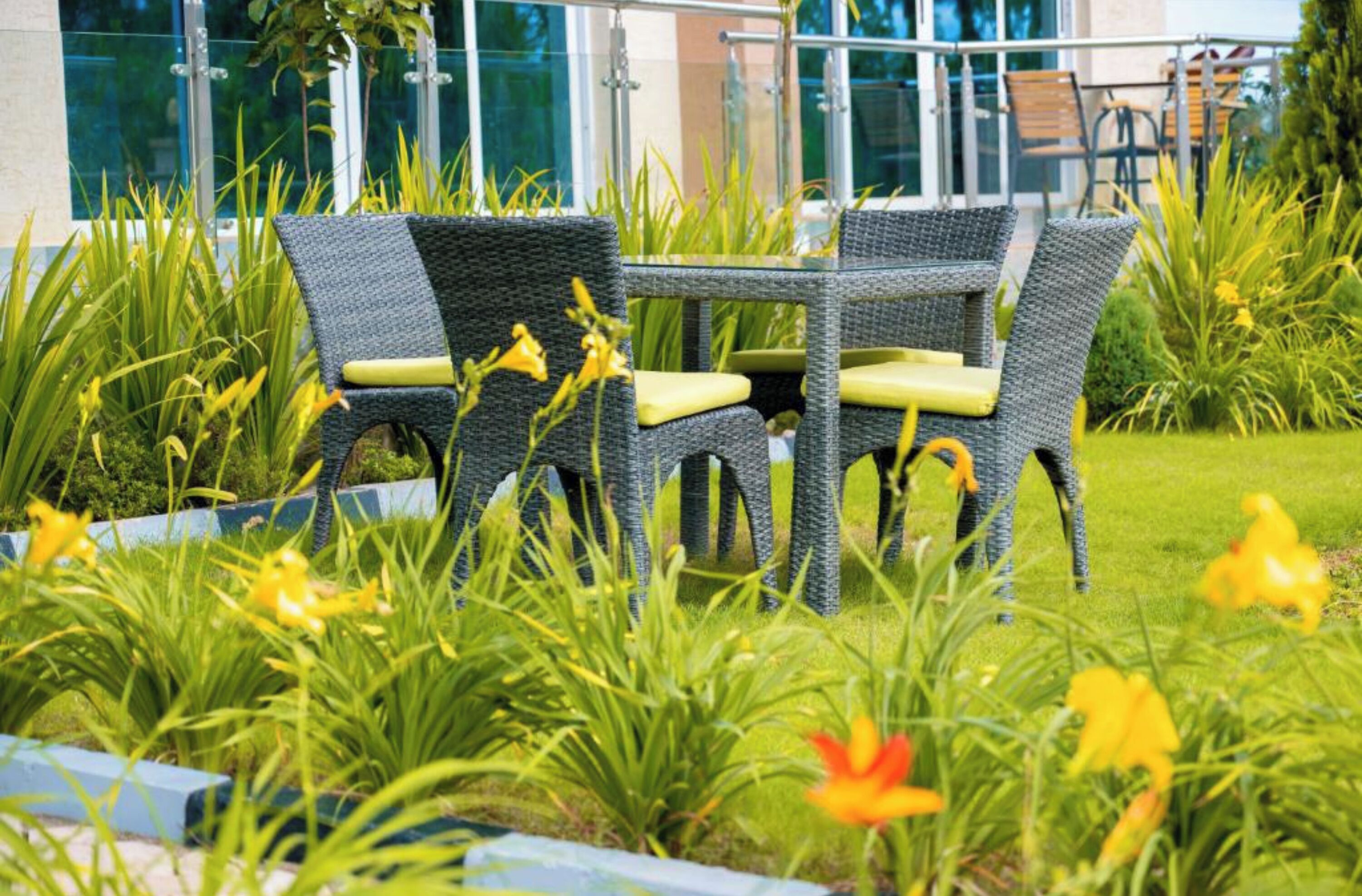 Un patio con mobiliario de mimbre y cojines amarillos, rodeado de abundante follaje y flores amarillas.