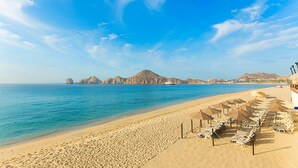 Una playa con filas de tumbonas y sombrillas, un mar azul cristalino y montañas a lo lejos.
