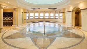 Una piscina interior amplia con una fuente central, rodeada por un suelo de baldosas en forma de círculo, y grandes ventanales a lo largo de las paredes.