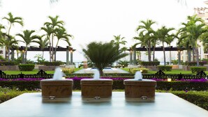 Una fuente de tres niveles en un jardín ajardinado con palmeras y una piscina al fondo.