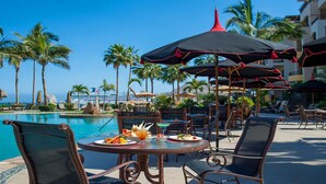 Un área de comedor junto a la piscina con una mesa lista para dos personas, rodeada de sillas y sombrillas.