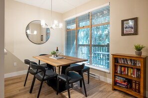 Un comedor con mesa de madera, sillas negras, una lámpara de araña, una estantería y un amplio ventanal con persianas.