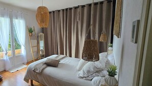 Een slaapkamer met een grote houten garderobe, een bed met witte lakens, een hangende rotanlamp en uitzicht op groen door dunne gordijnen.
