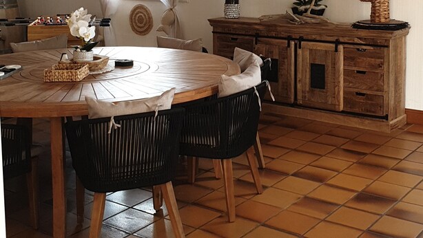 Een eetgedeelte met een ronde houten tafel, zwarte stoelen, een geweven hanglamp en een houten kast met lades.