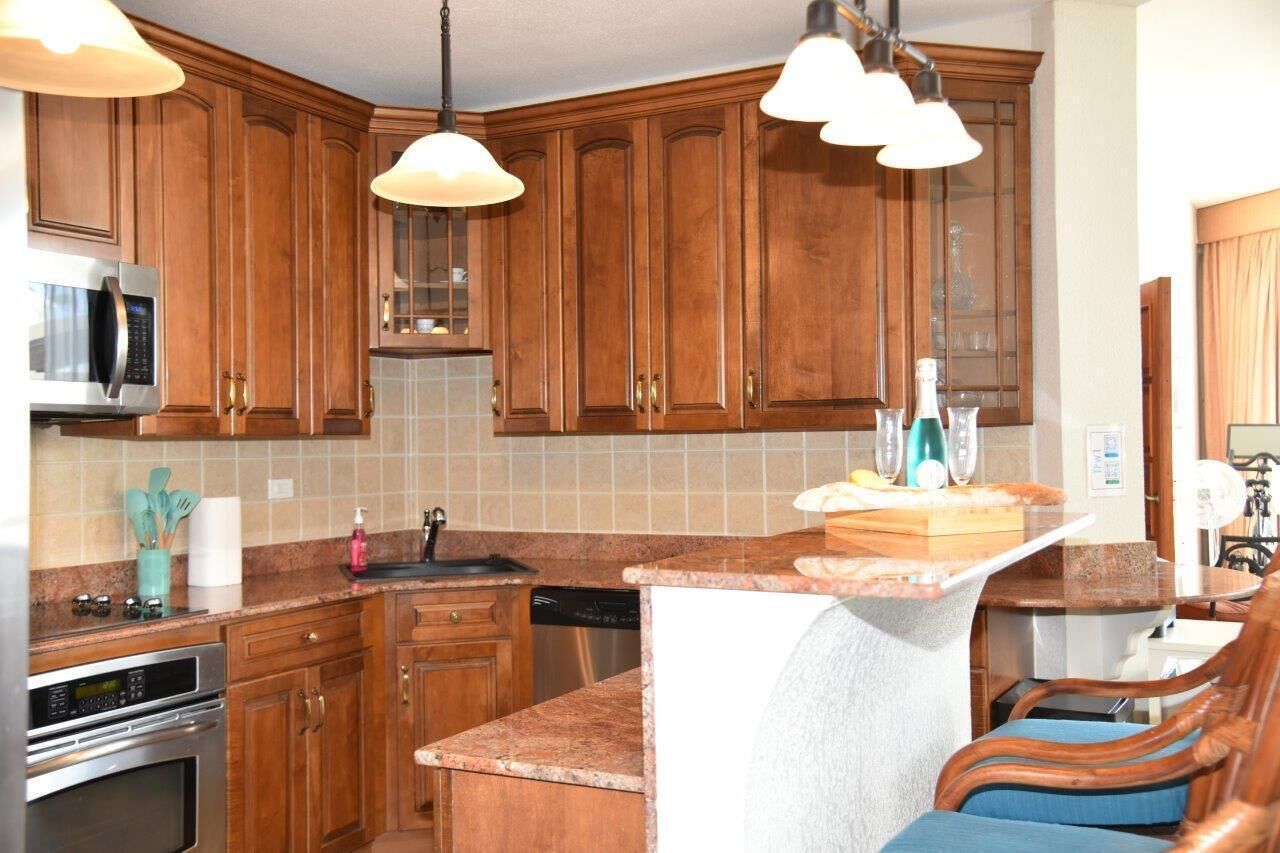 Una cocina con armarios de madera, microondas, horno y lavavajillas. Hay una pequeña isla con un fregadero y una silla.