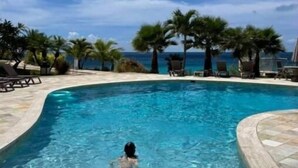 Une piscine à l’eau d’un bleu cristallin, entourée de chaises longues et de palmiers, offrant une vue sur l’océan et un ciel dégagé.