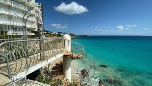 Un hôtel en bord de mer offrant une vue dégagée sur la mer turquoise, un escalier en pierre menant à un balcon, et un bâtiment blanc doté de nombreux balcons.