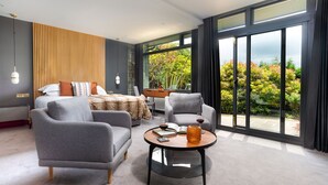 Ein modernes Schlafzimmer mit einem Bett, zwei Sesseln, einem kleinen Tisch und einem großen Fenster mit Blick auf Grünflächen.