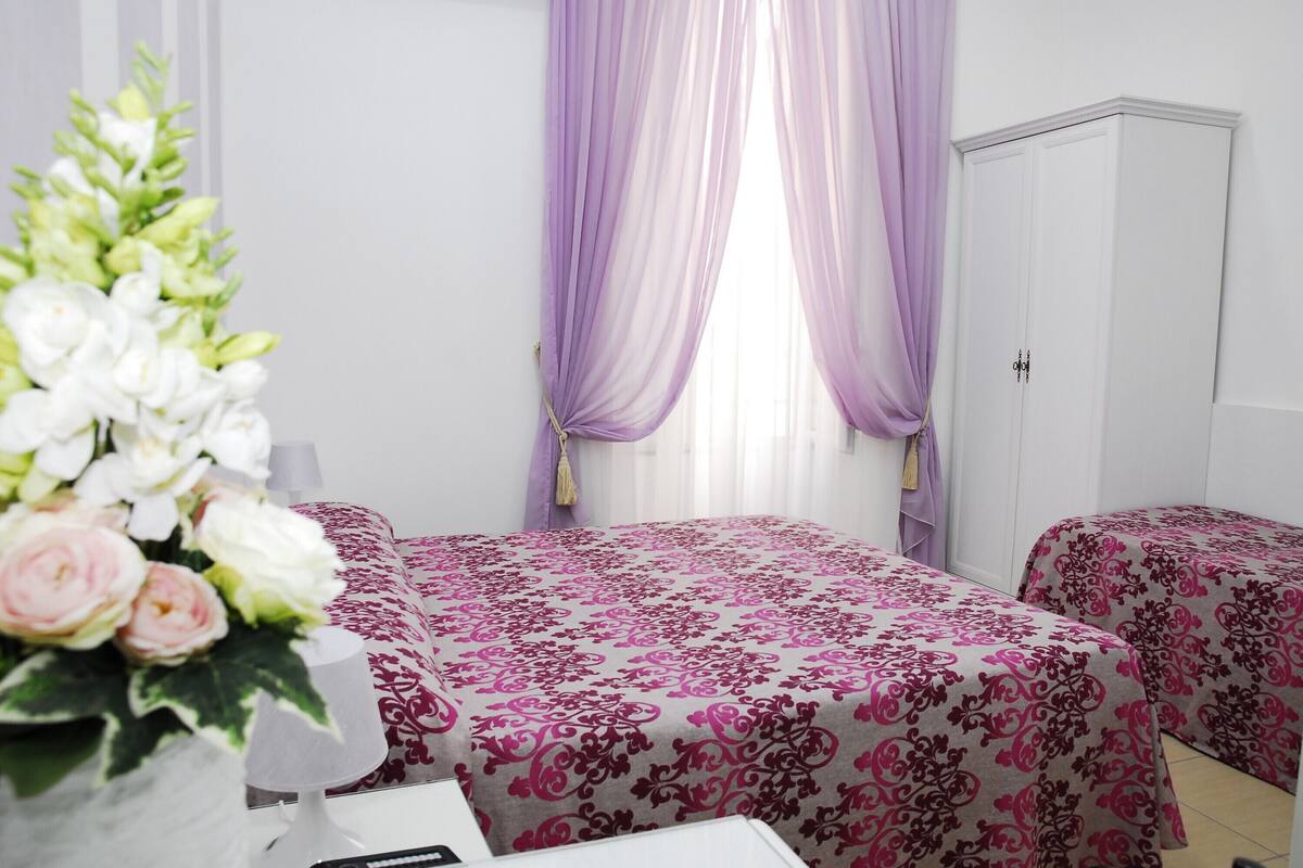 A hotel room with two beds, a white wardrobe, a window with purple curtains, and a vase of flowers.