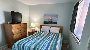 Un dormitorio con una cama, un aparador, un televisor sobre el aparador, una lámpara, un cuadro de un barco en la pared y una ventana.