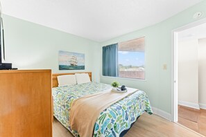 A bedroom with a bed, a painting, a window with curtains, and a door. - Carolina Reef 202 - 1 Bedroom Ocean Front - Pool & Hot Tub (North Myrtle Beach)