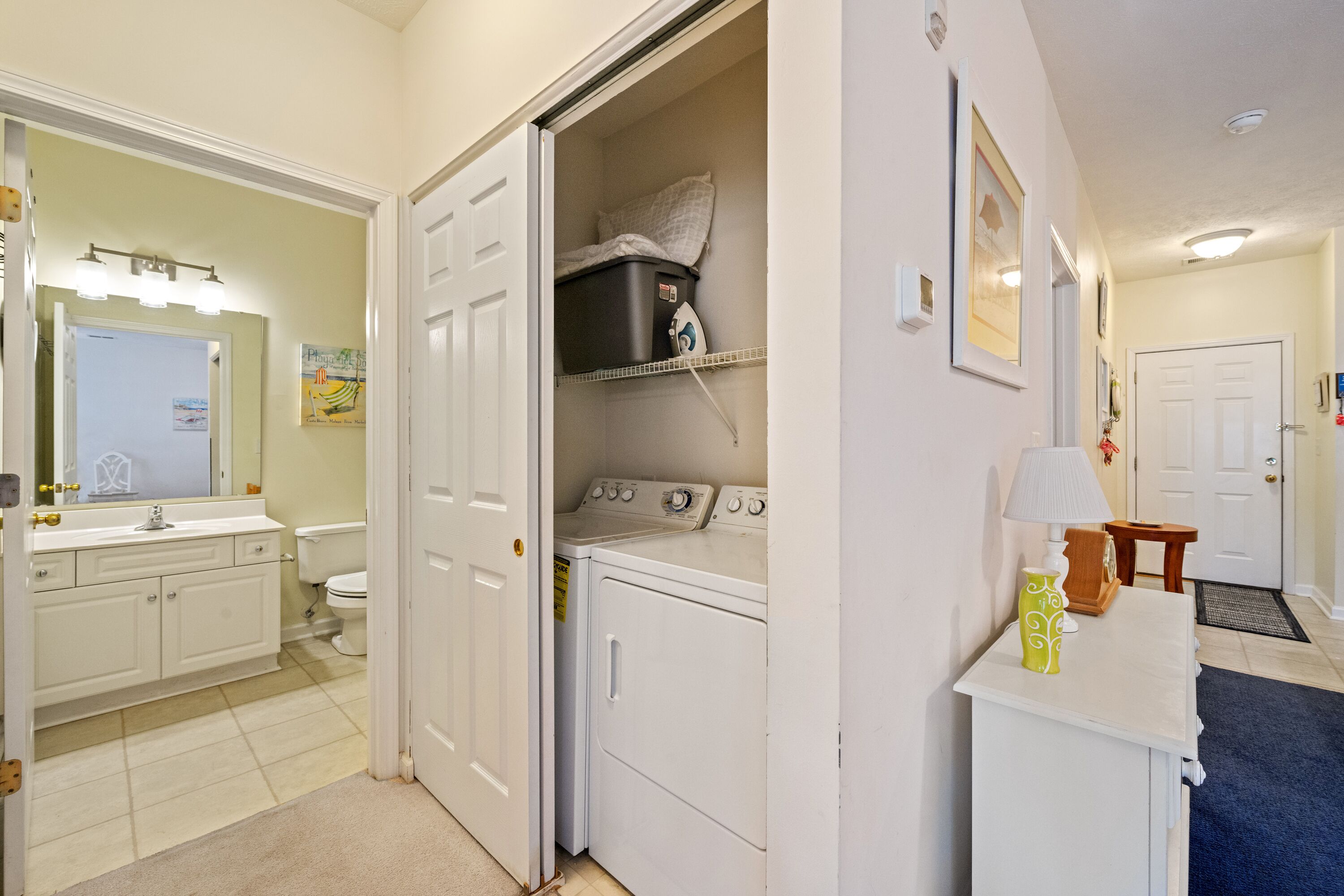 A laundry room with a washer and dryer, a small shelf with a pillow, and a door leading to a bathroom.