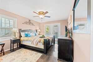 Une chambre à coucher comprenant un lit, des tables de chevet, une commode, un ventilateur de plafond et une fenêtre avec des stores.