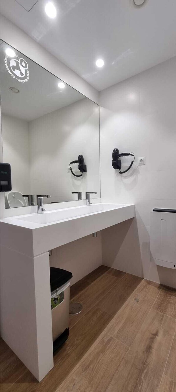 A modern bathroom with a white sink, mirror, and two hair dryers mounted on the wall. - Cantabria Surf Hostel (Ribamontan al Mar)