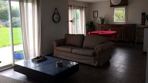 A living room with a sofa, a coffee table, a red table, and a view of a garden through the sliding glass door. - House 6 persons near Port de Doëlan / 2 stars tourism (Clohars-Carnoët)