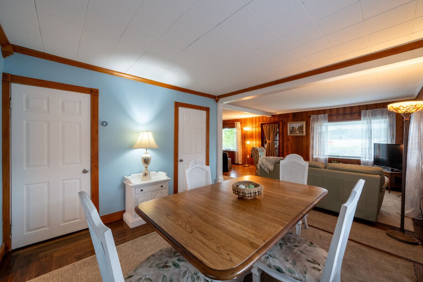 A cozy living room with a wooden dining table, white chairs, and a sofa. There is a lamp on a side table, a television, and a window with curtains.