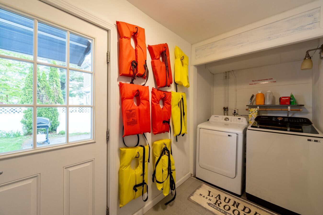 A laundry room with a washing machine and dryer, a window with a view of greenery, and life jackets hanging on the wall.