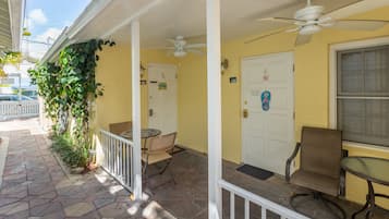 A covered porch with a white railing, a table, and two chairs. There is a door with a number sign, a white fence, and a window with blinds.