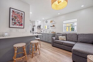 A modern kitchen with a dining area, a grey sofa, and a wicker bar stool. - The Dancing Rooster: Newly Renovated Key West Retreat, One Block to Duval! (Key West)