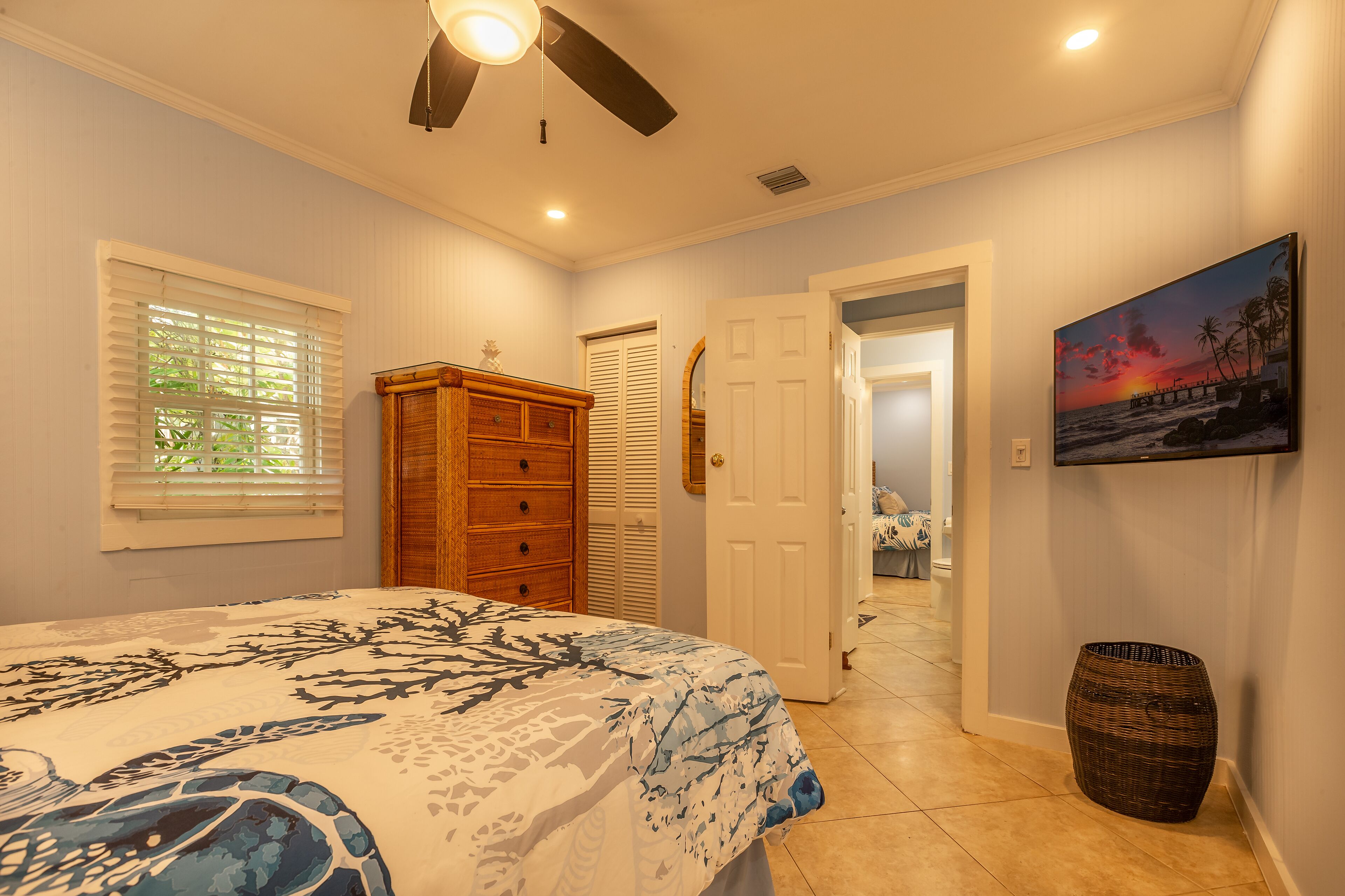 A bedroom with a bed, a ceiling fan, a window with blinds, a wooden dresser, a door, and a painting on the wall.