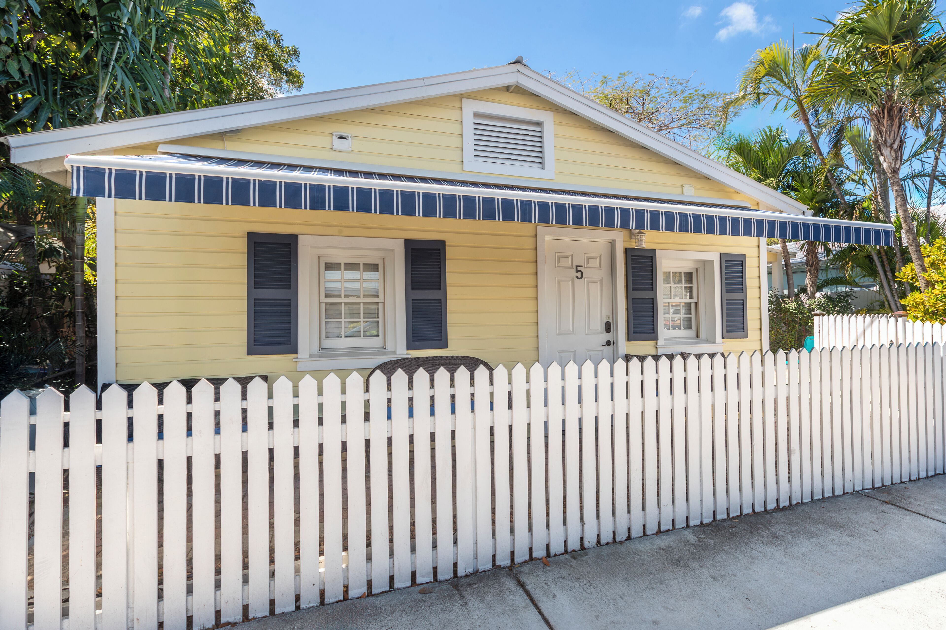A yellow house with a white picket fence, a number '5' on the door, and a small porch.