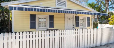A yellow house with a white picket fence, a number '5' on the door, and a small porch.