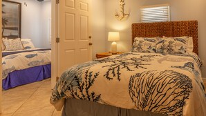 Quarto com cama, mesa de cabeceira, luminária, ventilador de teto, porta e um enfeite fixado na parede.