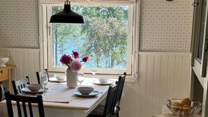 Un coin repas avec une table dressée pour quatre personnes, des chaises, une fenêtre donnant sur les arbres, une horloge au mur et un comptoir de cuisine sur lequel se trouve un bol de citrons.